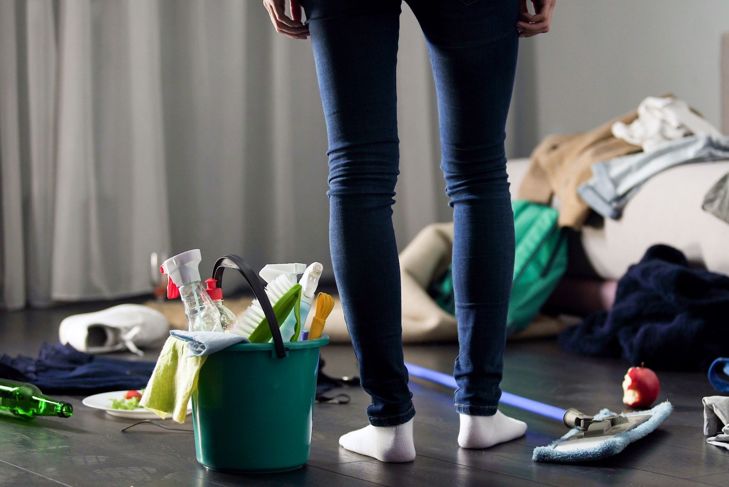 Woman stands in front of messy room with cleaning supplies
