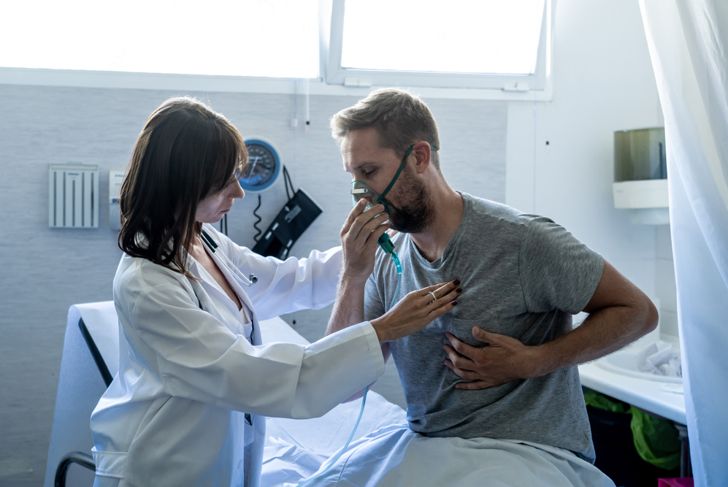 Young sick man patient wearing oxygen mask while female doctor listening his chest with stethoscope