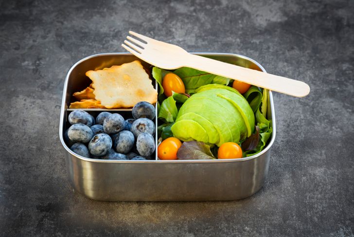 Lunch box with sliced ​​avocado, yellow tomatoes, crackers, blueberries and green salad