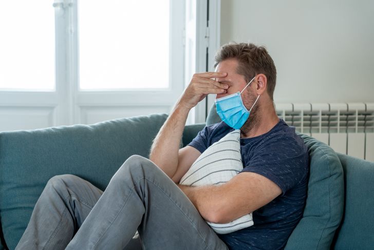 Sad man wearing protective face mask at home, sofa in living room, tired