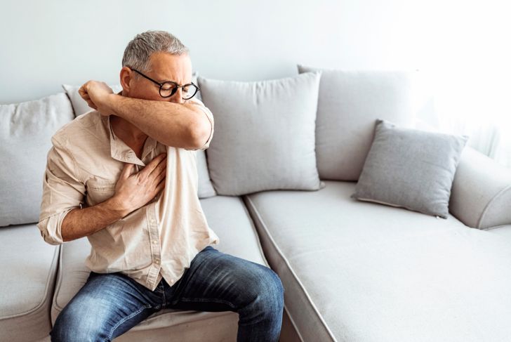 man coughs into his elbow while sitting on the couch at home