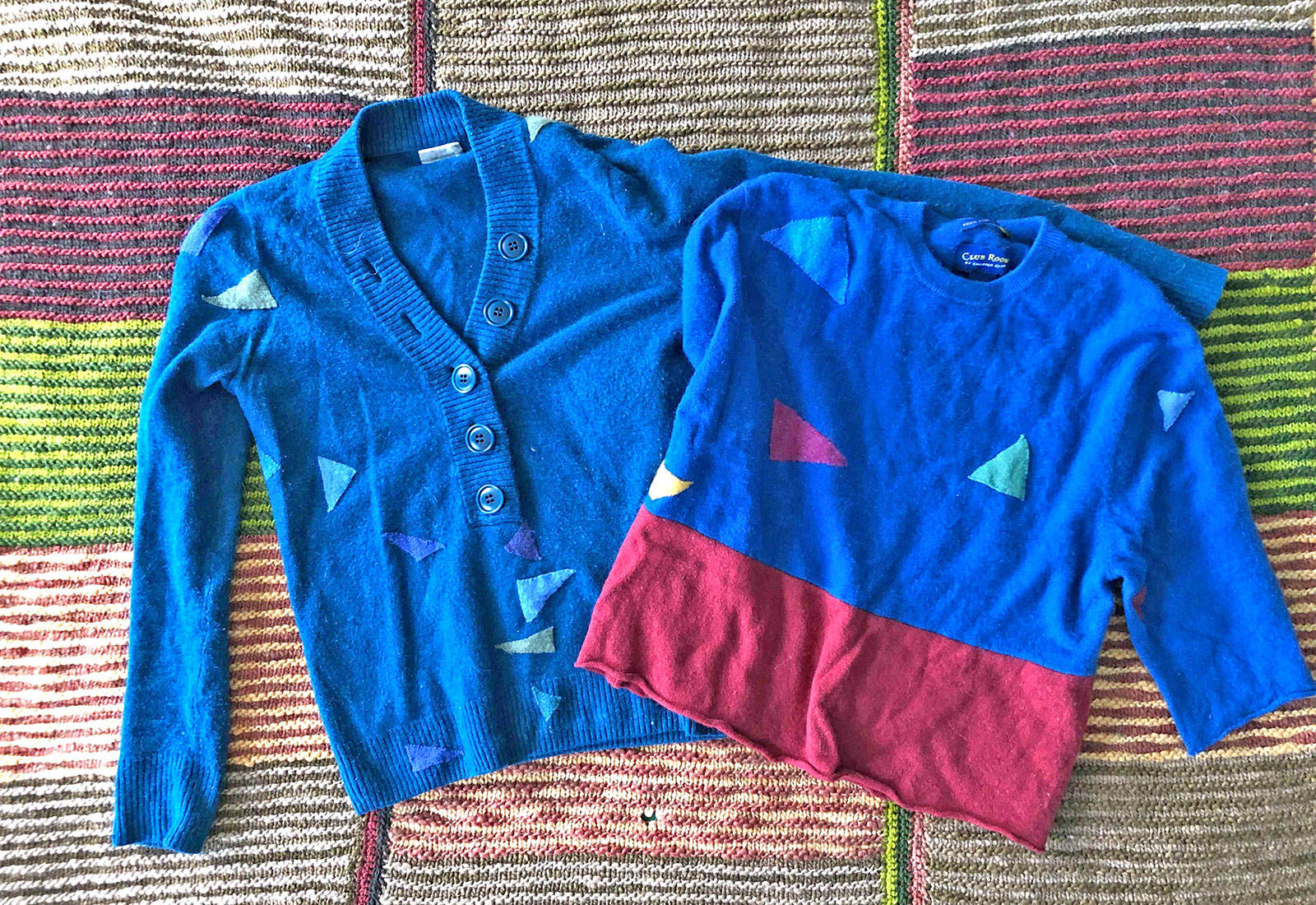 Two blue sweaters with triangular patches on top of a colorful green and red blanket
