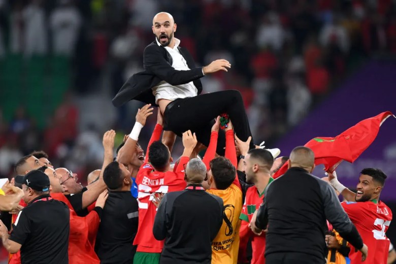 A man in a suit and white shirt is lifted high into the air by a team of men in football uniforms in the red and green of morocco.